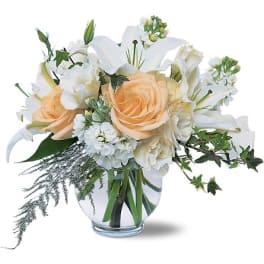 Low glass vase of peach roses and white lilies with white accent flowers and trailing greenery