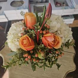 Compact arrangement of orange roses, tulip buds, white hydrangeas, and berries on a small table.