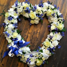 Heart-shaped wreath of white flowers with yellow roses, blue blooms, and a blue and white ribbon bow