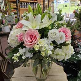 Pink roses, white lilies, and white hydrangeas arranged in a clear glass vase on a small wooden table