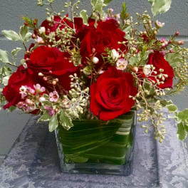 Red rose arrangement with pink filler blooms in a leaf-wrapped glass cube vase