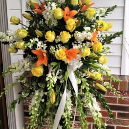 Tall standing spray with yellow roses, orange lilies, white flowers, and trailing green stems on an easel.