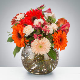 Round glass bowl vase of orange and red gerbera daisies with cream carnations and pink blooms.