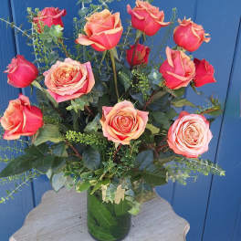 Tall arrangement of coral and orange roses in a clear glass vase on a small wooden table