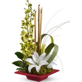 White lilies in a red square vase with green orchids and bamboo sticks
