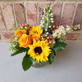 Low glass vase of yellow sunflowers, orange roses, and mixed accent blooms.