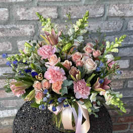 Mixed arrangement of pink roses, carnations, lilies, and blue flowers in a clear vase with a pink ribbon.