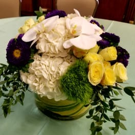 Bouquet of white hydrangeas, yellow roses, and purple flowers in a glass vase