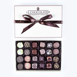 Rectangular box of assorted gourmet chocolates with lid and brown ribbon beside open tray