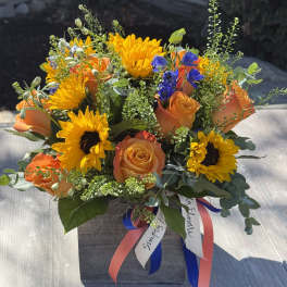 Square wooden box arrangement with yellow sunflowers, orange roses, and blue accent flowers with ribbons