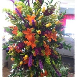 Tall standing spray of orange lilies, yellow roses, and purple flowers on a metal easel