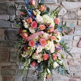 Tall standing spray of peach and white roses with blue accents on a metal easel against a stone wall.