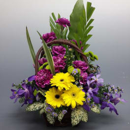 Basket arrangement with yellow gerbera daisies, purple flowers, and tropical leaves