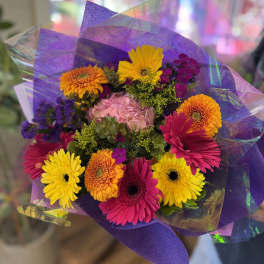 Bright hand-tied bouquet of pink, yellow, and orange gerbera daisies with mixed blooms in purple wrap