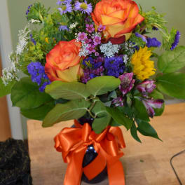 Bouquet of orange roses, purple flowers, and yellow blooms in a dark vase with an orange ribbon