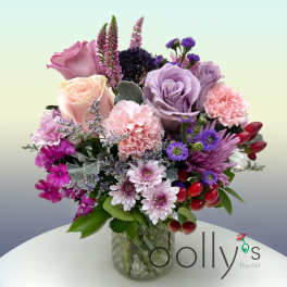 Mixed bouquet of pink and lavender roses, carnations, and purple blooms in a clear glass vase.