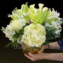 Low arrangement of white and pale green hydrangeas, roses, lilies, and an orchid in an amber glass cube vase.
