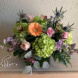 Low arrangement of green hydrangeas, peach gerbera, pink roses, orchids in a glass vase beside pearl beads