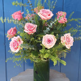 Tall arrangement of pink roses with airy filler in a clear glass vase on a small wooden table