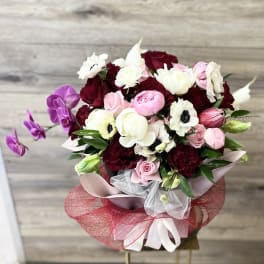 Round bouquet of red, pink, and white blooms with purple orchids and decorative red and white wrapping.