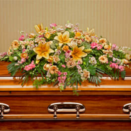 Casket spray of yellow lilies with pink and peach flowers on a polished wood casket.
