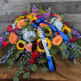 Colorful casket spray with sunflowers, roses, hydrangeas and a blue ribbon reading Loving Husband