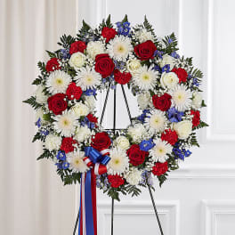 Circular funeral wreath of red, white, and blue flowers on a black stand