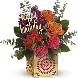 Birthday arrangement with orange roses, pink carnations, and purple flowers in a patterned cube vase.