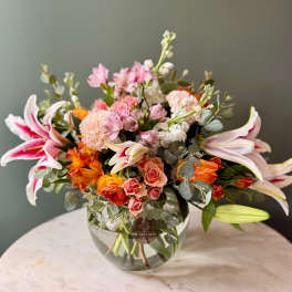 Mixed arrangement of pink lilies, roses, and orange blooms in a round glass vase
