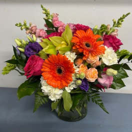 Mixed bouquet with orange gerbera daisies and pink, purple, and white flowers in a glass vase