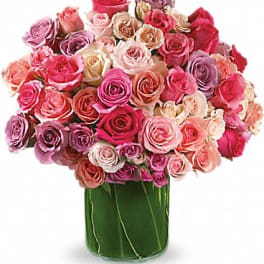 Lush bouquet of pink, peach, and lavender roses in a clear cylinder vase wrapped with green leaves.