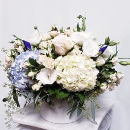 Low arrangement of white and blue hydrangeas, roses, and lisianthus in a round white container