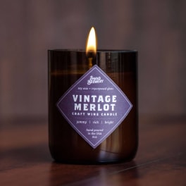 Lit wine-themed candle in a dark glass jar with a purple label on a wood surface