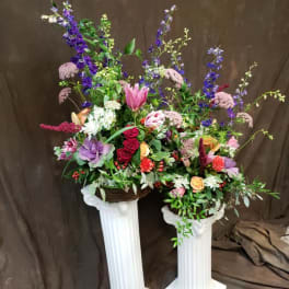 Two tall floral arrangements in white pedestal stands with pink, purple, and red blooms.