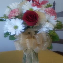 Mixed bouquet of red roses, white daisies, and pink carnations in a glass vase with gold bow and red butterfly pick