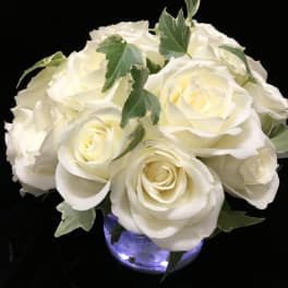 Low arrangement of white roses in a round glass vase with soft interior lights.