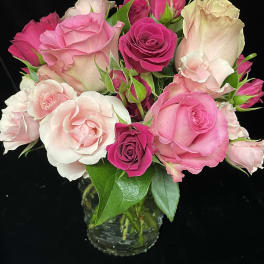 Pink and blush roses arranged tightly in a small clear glass vase against a black background