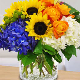 Bouquet of sunflowers, orange roses, blue hydrangeas, and white blooms in a glass vase