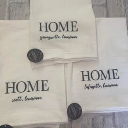 Stacked white towels printed with "HOME" and Louisiana town names