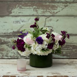 Purple and white floral arrangement in a dark green vase with a small candle nearby
