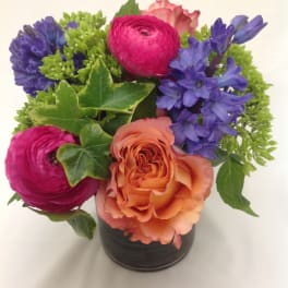 Compact arrangement of peach rose, pink ranunculus, purple hyacinths and green blooms in a small vase with ivy