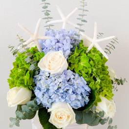 Blue hydrangeas and white roses arranged with starfish accents in a white container