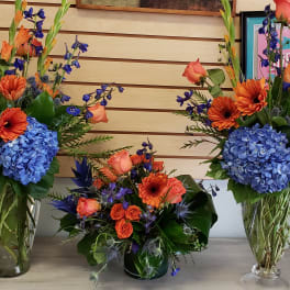 Three floral arrangements with orange, blue, and purple flowers in glass vases