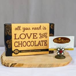 Box of assorted chocolates with love-themed text on a wooden board beside a floral card holder.