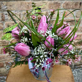 Pink tulip arrangement with white filler flowers, grass loops, and decorative butterflies in a metal pail.
