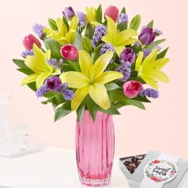 Yellow lilies and pink tulips in a pink vase with a candy box beside it
