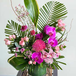 Pink orchids and roses arranged in a glass vase with large tropical leaves