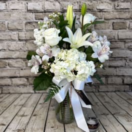 Tall white flower arrangement with lilies, roses, and hydrangeas in a glass vase with a ribbon bow.