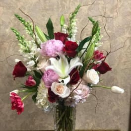 Tall mixed arrangement of pink peonies, red and white roses, lilies and tulips in a clear glass vase