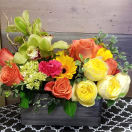 Low wood box arrangement with orange roses, yellow blooms, green orchids, and a pink carnation.